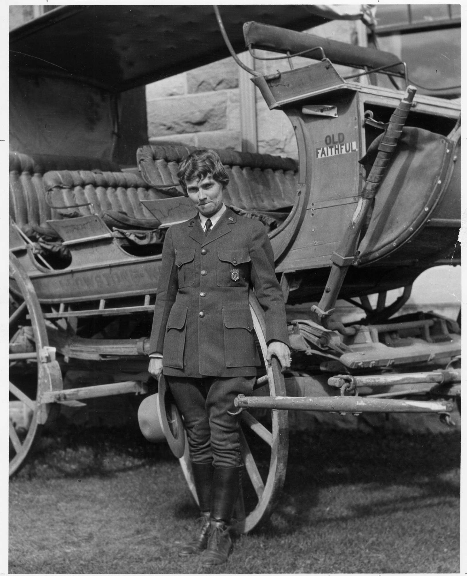 Herma Albertson poses in front of a stagecoach while wearing the NPS uniform with shield-shaped badge and holding a broad brim hat.
